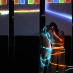 Two performers dressed in white trousers and vests with long hair stand side-by-side in a darkened room with strips of flexible lighting wound round their bodies one is bright blue and the other orange, some coloured light squares are big above and behind them