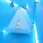 A white suspended tent in a blue-lit room, with text projected onto the floor and neon lights illuminating the walls.