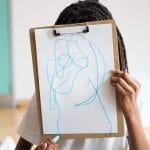 A school child with long dreadlocks and white shirt hides their face behind a clipboard holding a piece of A4 paper into which they have drawn a self portrait with a blue pen