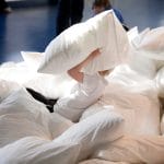 A school child plays in a big pile pf bright white pillows, catching one as it hides their face, other children in the background watch