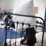 One school child kneels on an old fashioned single bed, with a decorative railed headboard, they hide their face with a pillow, and other children are in the background
