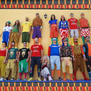 a group of seventeen performers in brightly pattered and colourful, mismatched clothes and all wearing sunglasses form a two-level human tower in front of a yellow wall.