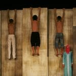 five performers hang by their hands from a tall wall which they face, they wear trousers only apart from one who also wears a red shirt while a sixth performer, dressed in a light blue outfit and a pink headscarf, stands below looking at the camera, we can only see their eyes from in the headscarf