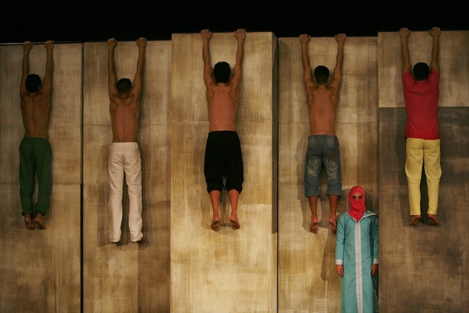 five performers hang by their hands from a tall wall which they face, they wear trousers only apart from one who also wears a red shirt while a sixth performer, dressed in a light blue outfit and a pink headscarf, stands below looking at the camera, we can only see their eyes from in the headscarf
