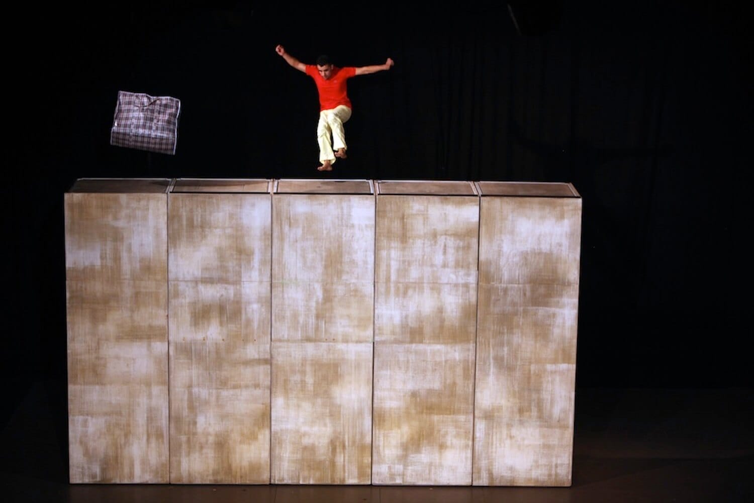 Person jumping off a block of large boxes on a stage with a checkered bag hung in the air.