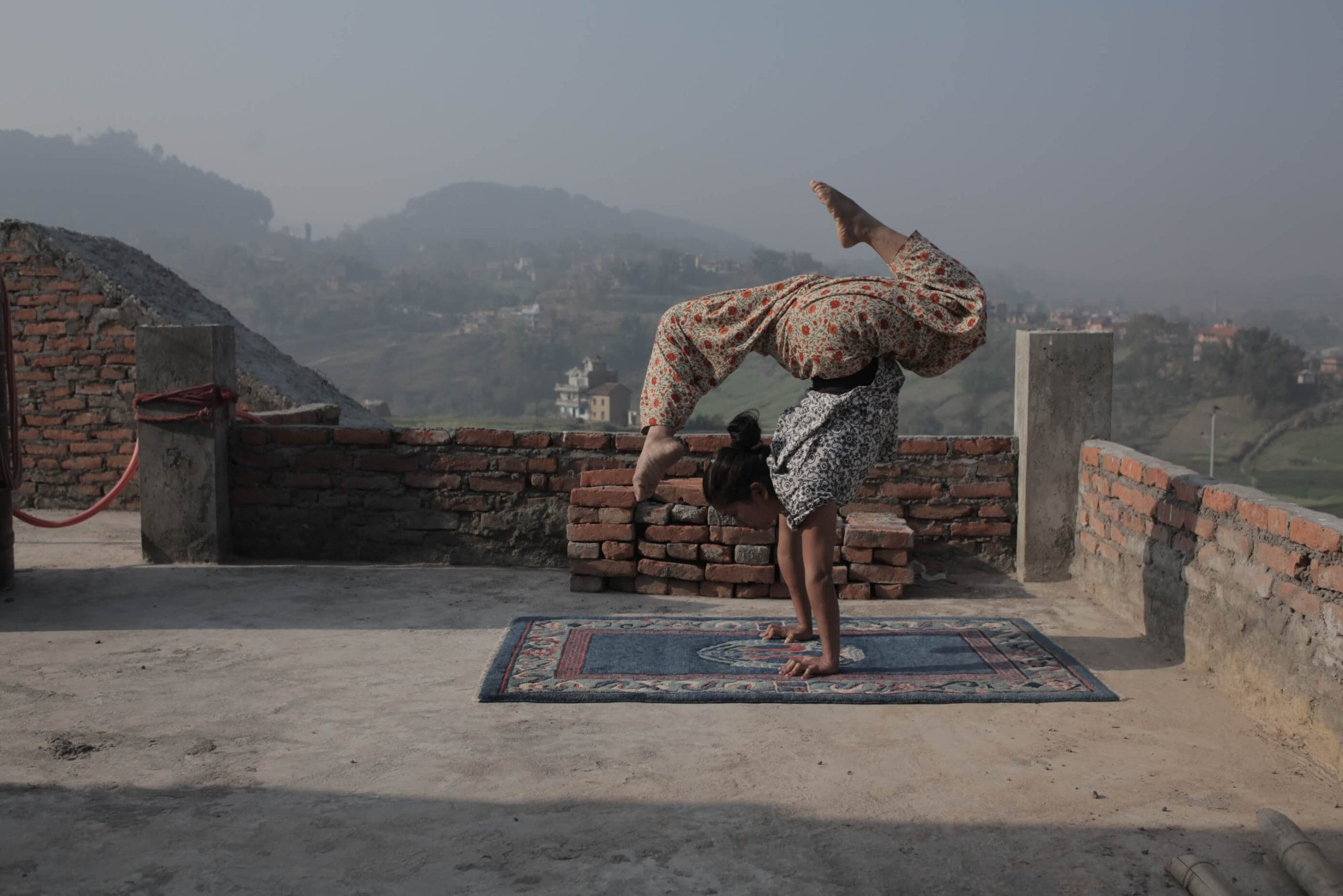 Person performing an outdoor handstand on a small patterned rug, on a rooftop, with a scenic hilly and hazy landscape in the background, they cast a shadow on the rooftop and wall