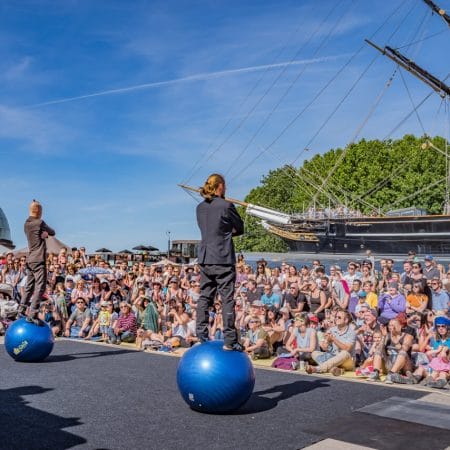 two performers each stand on a blue exercise ball place on a floor level, outdoor stage, one has tied back, brown hair and a beard, wearing a dark trouser suit, the other has short brown hair, black jeans and sweater, they face a large audience seated in bright blue sunshine, a docked boat is visible in the background
