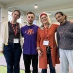 four people stand happily in a row with their arms round each other, smiling at the camera, in a school gym, two are Parkour performers and two are managing the Parkour workshop