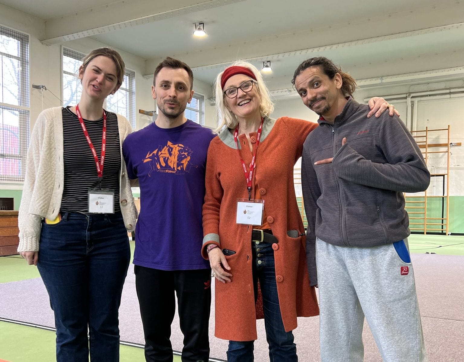 four people stand happily in a row with their arms round each other, smiling at the camera, in a school gym, two are Parkour performers and two are managing the Parkour workshop