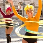 Two performers in vibrant yellow and red costumes and hair wraps, twirling colourful hula hoops in front of a historic building.