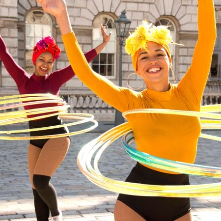 Two performers in vibrant yellow and red costumes and hair wraps, twirling colourful hula hoops in front of a historic building.
