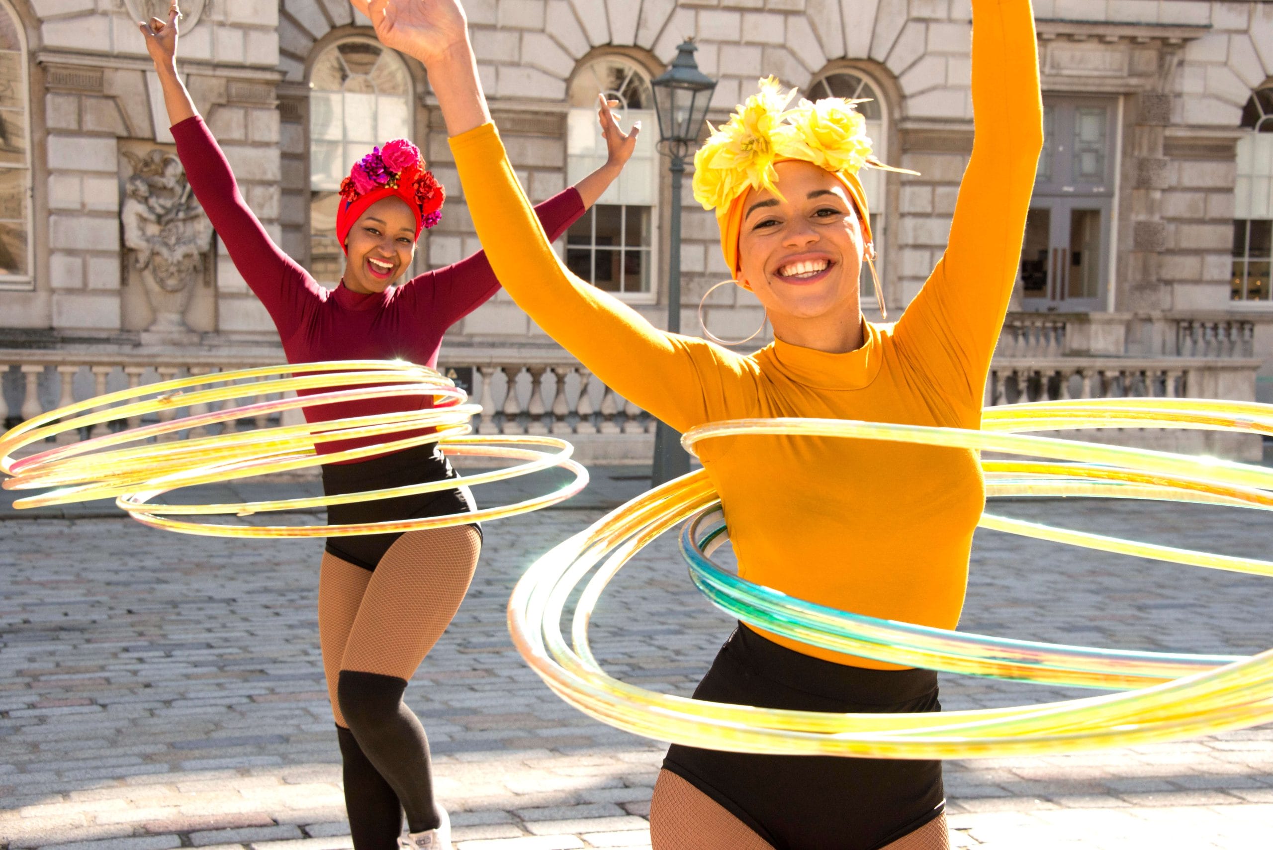 Two performers in vibrant yellow and red costumes and hair wraps, twirling colourful hula hoops in front of a historic building.