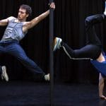 two performers are in an indoor rehearsal room, both connected to a Chinese pole, one in a grey vest and blue jeans with a dark beard holds on with one hand and braces their body with one foot, the other wearing a blue t-shirt and tracksuit bottoms with a read hat does a handstand bracing themself agains the pole with one foot