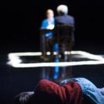 A young child with long blonde hair wearing a blue skirt and red top, lies in the foreground looking at an adult couple sitting at a table, one has short grey hair and a dark blue suit, the other has a blue robe over a white dress and long blonde hair, their table is surrounded by a white chalked square on the ground