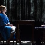 A young child with long blonde hair wearing a blue patterned skirt and red top, walks beside an adult couple who are sitting at a table, one has short grey hair and a dark blue trouser suit, the other has a blue robe over a pale dress and long blonde hair, their table is surrounded by a white chalked square on the ground