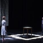 An adult couple are facing each other across a table, one has a black and white dress and long blonde hair tied up and is barefoot, the other has short grey hair and a dark blue trouser suit and pale shirt, the table is surrounded by a white chalked square on the ground