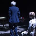 An adult couple are facing away from the viewer, one has a black and white dress and long blonde hair tied up and is sitting on a chair, the other has short grey hair and a dark blue suit, a table in the background is surrounded by a white chalked square on the ground