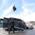 three performers stand in front of an open backed dark van on a floor level outdoor stage, the sky is blue and industrial looking buildings are in the distance, the performer are dressed in dark trouser suits and two are propelling the third up in the air from a teeterboard