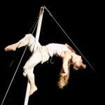 a performer with long blonde hair and dressed in cream trousers and a vest grips on a moving pole with only their legs, their arms and head are flung back and spotlit against a dark background