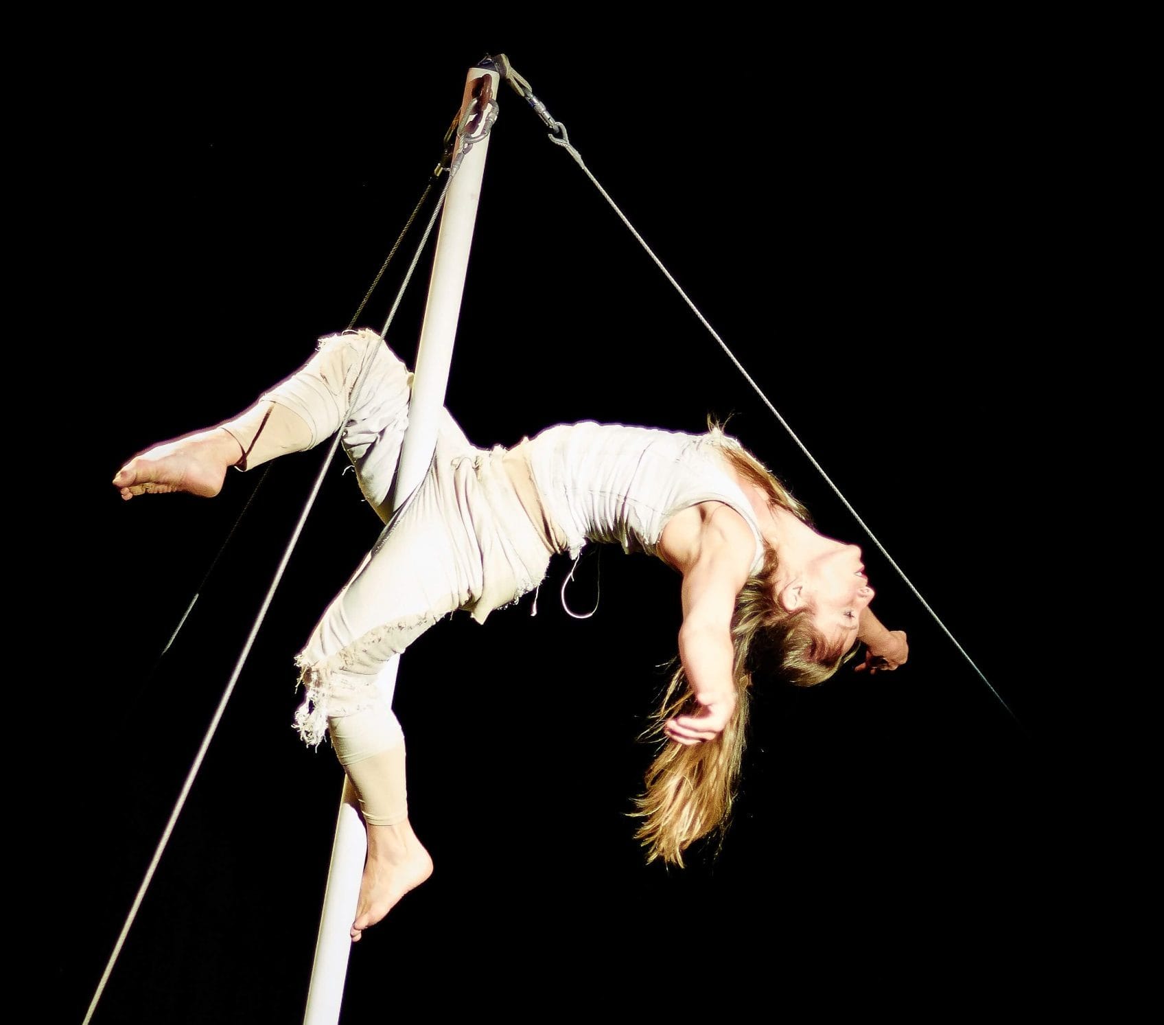 a performer with long blonde hair and dressed in cream trousers and a vest grips on a moving pole with only their legs, their arms and head are flung back and spotlit against a dark background