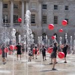 Performers juggle red balls and dance among fountains in front of a historic building.
