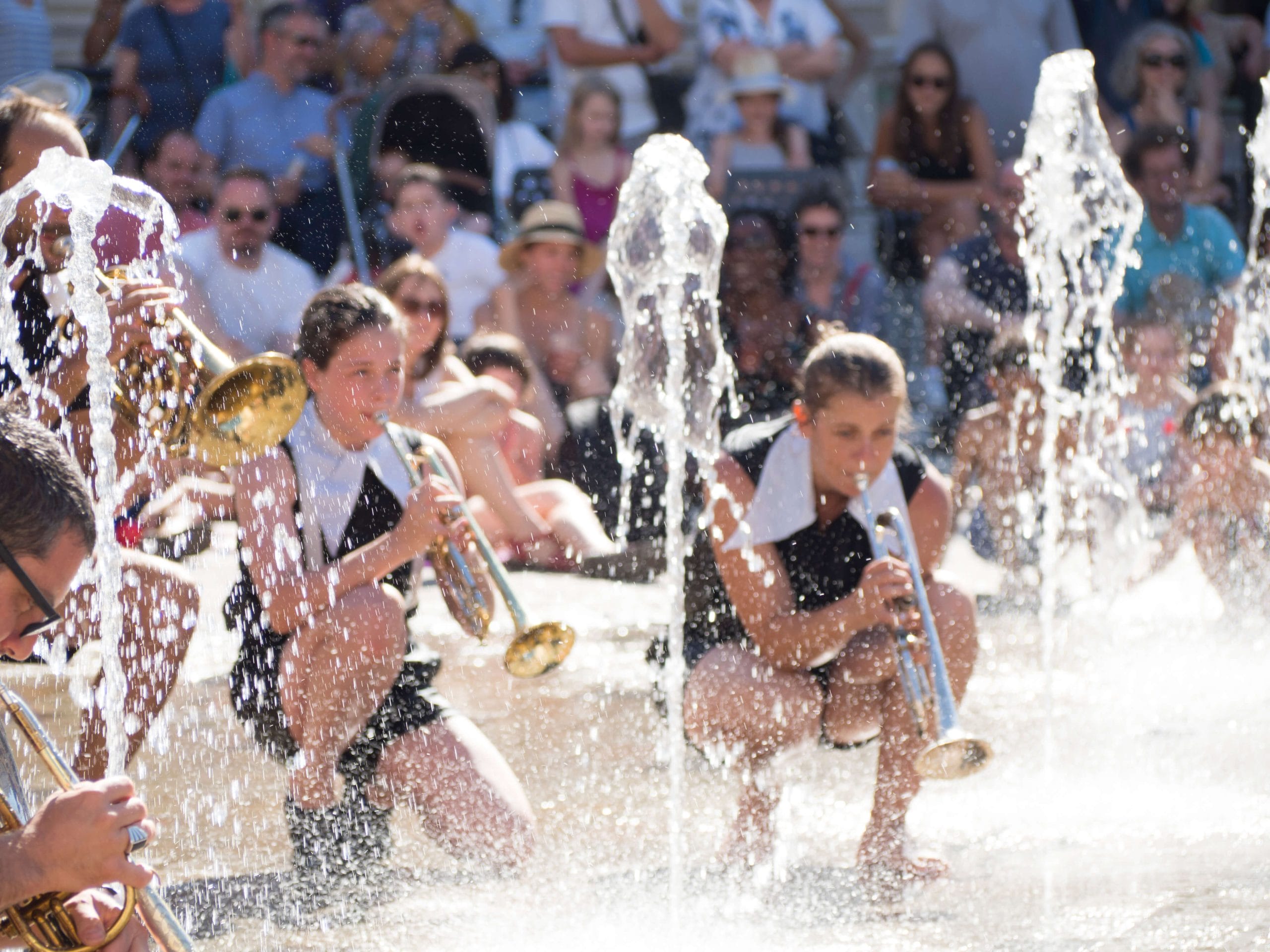 Musicians with trumpets perform among fountains on a sunny day, with a crowd watching in the background.