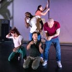 five performers move in a close group around a Chinese pole in a indoor rehearsal room, their clothes are casual and colourful and they appear to be in their own worlds with hands and arms creating movement