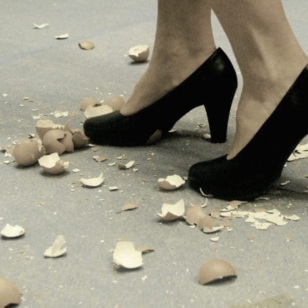 A pair of feet in black high heels walking delicately amongst scattered broken eggshells on the ground