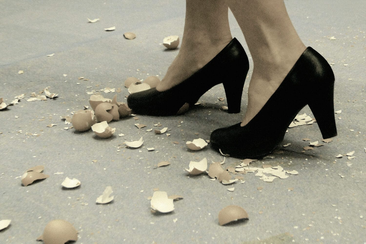 A pair of feet in black high heels walking delicately amongst scattered broken eggshells on the ground
