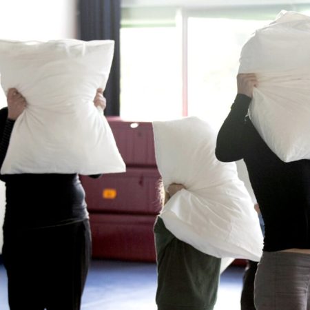 A group of five school children hide their faces with pillows, standing against a room with a wide backdrop of a window.