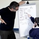 A school child points at a whiteboard with writing and drawings, while an adult dressed in black stands beside it, smiling at the board.