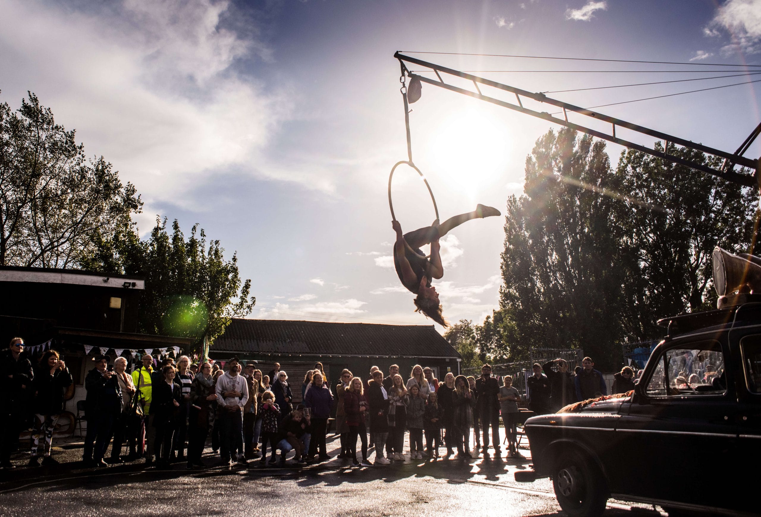 Aerial acrobat performing in a hoop above a crowd outdoors, silhouetted against a sunlit sky.