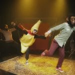 Three performers are on a stage: one in a monkey mask holding hands with another in a banana costume, while the third stands at the back of the row.