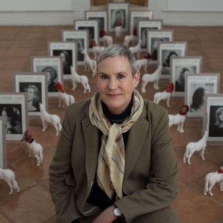 Person in a brown coat a patterned neck scarf with cropped grey hair and stud earrings surrounded by small white horse figurines, some wearing little headscarves and also seen are many different framed Country Life magazine covers.