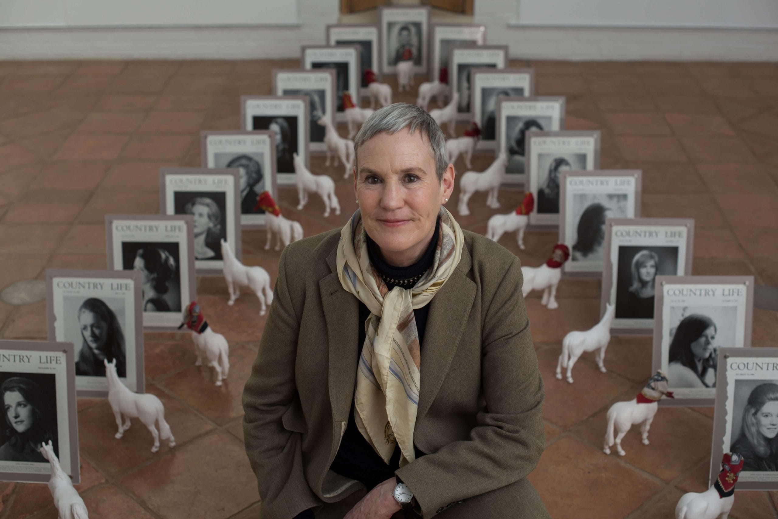 Person in a brown coat a patterned neck scarf with cropped grey hair and stud earrings surrounded by small white horse figurines, some wearing little headscarves and also seen are many different framed Country Life magazine covers.