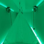 Green-lit room featuring a triangular suspended tent at the centre, illuminated by neon lights.