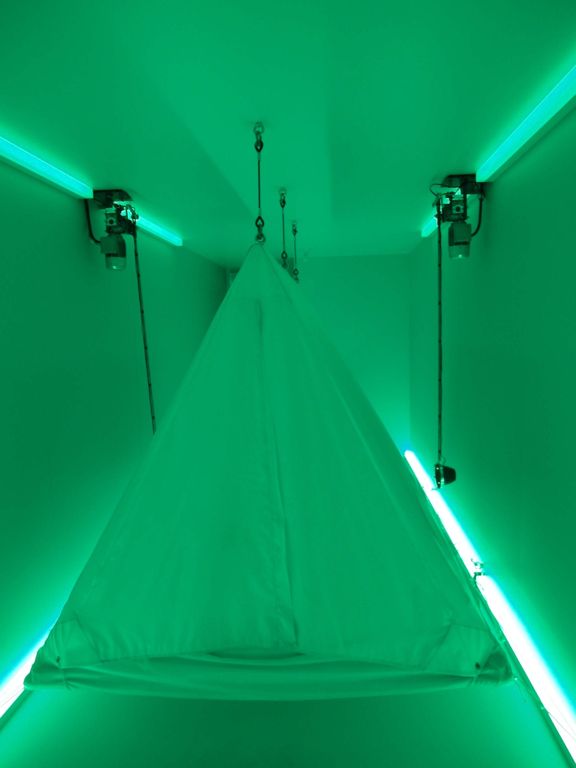Green-lit room featuring a triangular suspended tent at the centre, illuminated by neon lights.
