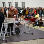 two performers stand on a floor level, outdoor stage, one has tied back, brown hair and a beard, wearing a dark trouser suit, the other has short brown hair, black jeans and a brown striped sweater, they are lifting a series of different types of weights while a large audience look on