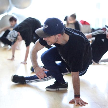 a performer is in the foreground dressed in a dark tracksuit and dark baseball hat, one hand and both feet are on the floor mid Parkour move with people in the background out of focus. Sasha Biloshisky.