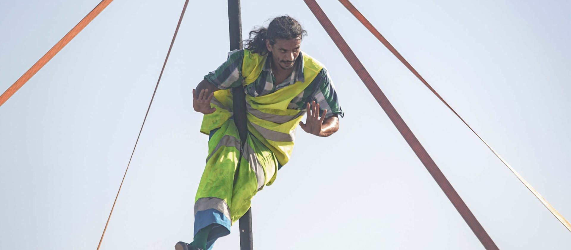 an acrobat dressed in hi-vis clothing with long dark hair and dark facial hair is balancing in the air on a Chinese pole set up outdoors against pale blue sky Taroo parkour circus show