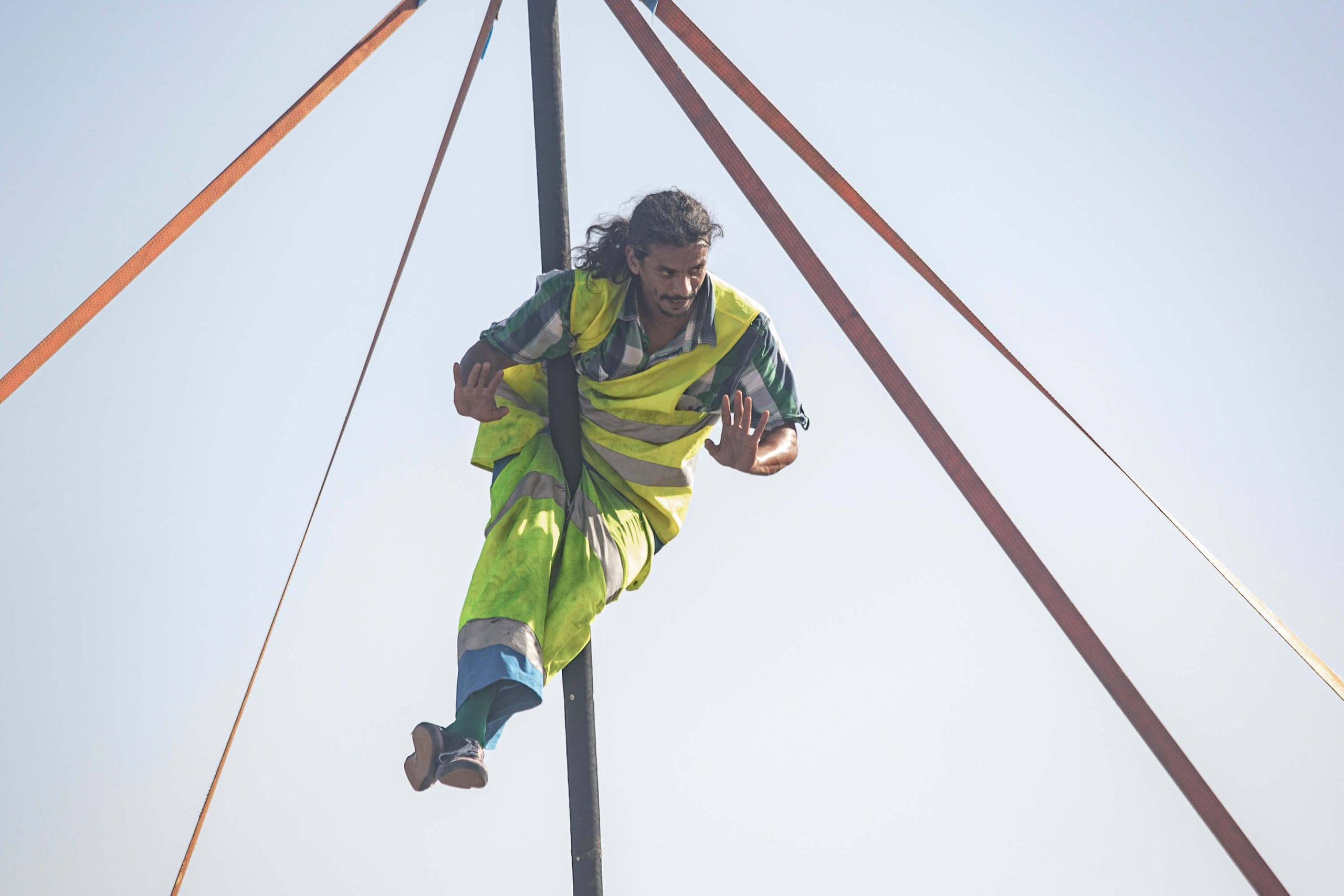 an acrobat dressed in hi-vis clothing with long dark hair and dark facial hair is balancing in the air on a Chinese pole set up outdoors against pale blue sky Taroo parkour circus show
