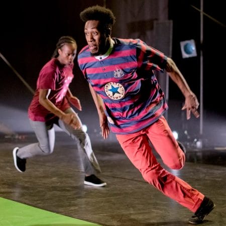 two performers run round an indoor rehearsal space, one has an afro hairstyle and wears red jeans and a striped t-shirt, the other has dreadlocks and grey jeans with a red t-shirt