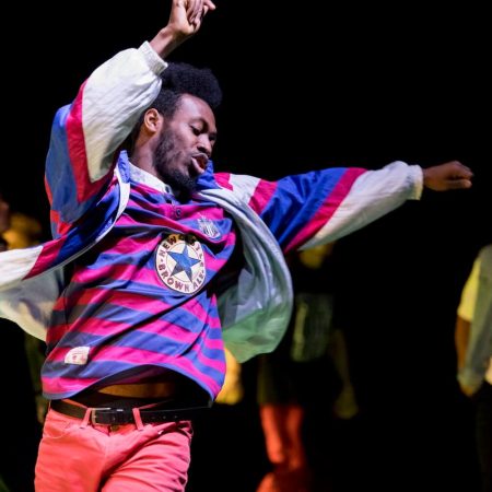 one performer with an afro dances in an indoor rehearsal space wearing red jeans, a striped t-shirt, and a red white and blue jacket, other performers are in the background slightly blurred