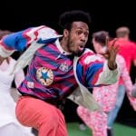 one performer with an afro dances in an indoor rehearsal space wearing red jeans, a striped t-shirt, and a red white and blue jacket, other performers are in the background slightly blurred, one dancer in a white wedding dress and another in a Union Jack suit