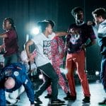 seven performers are all striking poses which suggest a big fight, they are spotlit from behind with other performers in the background, all in an indoor rehearsal space, everyone is wearing colourful casual clothing