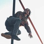 an acrobat dressed in blue jeans and a brown shirt with long dark hair and dark facial hair is balancing in the air on a Chinese pole set up outdoors against pale blue sky