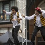 Two animated performers in back trousers, white shirts and yellow and black diamond pattered waistcoats stand side by side on an open air stage.