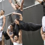 nine acrobats dressed in blacks, greys and whites form a three story human tower against a colourful backdrop