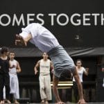 one acrobat performs a cartwheel with five acrobats watching and clapping behind, they're dressed in blacks, greys and whites