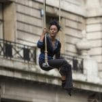 a performers in brown trousers and a blue and black shirt sits on an outdoor, high, swinging trapeze against a backdrop of grand stone buildings